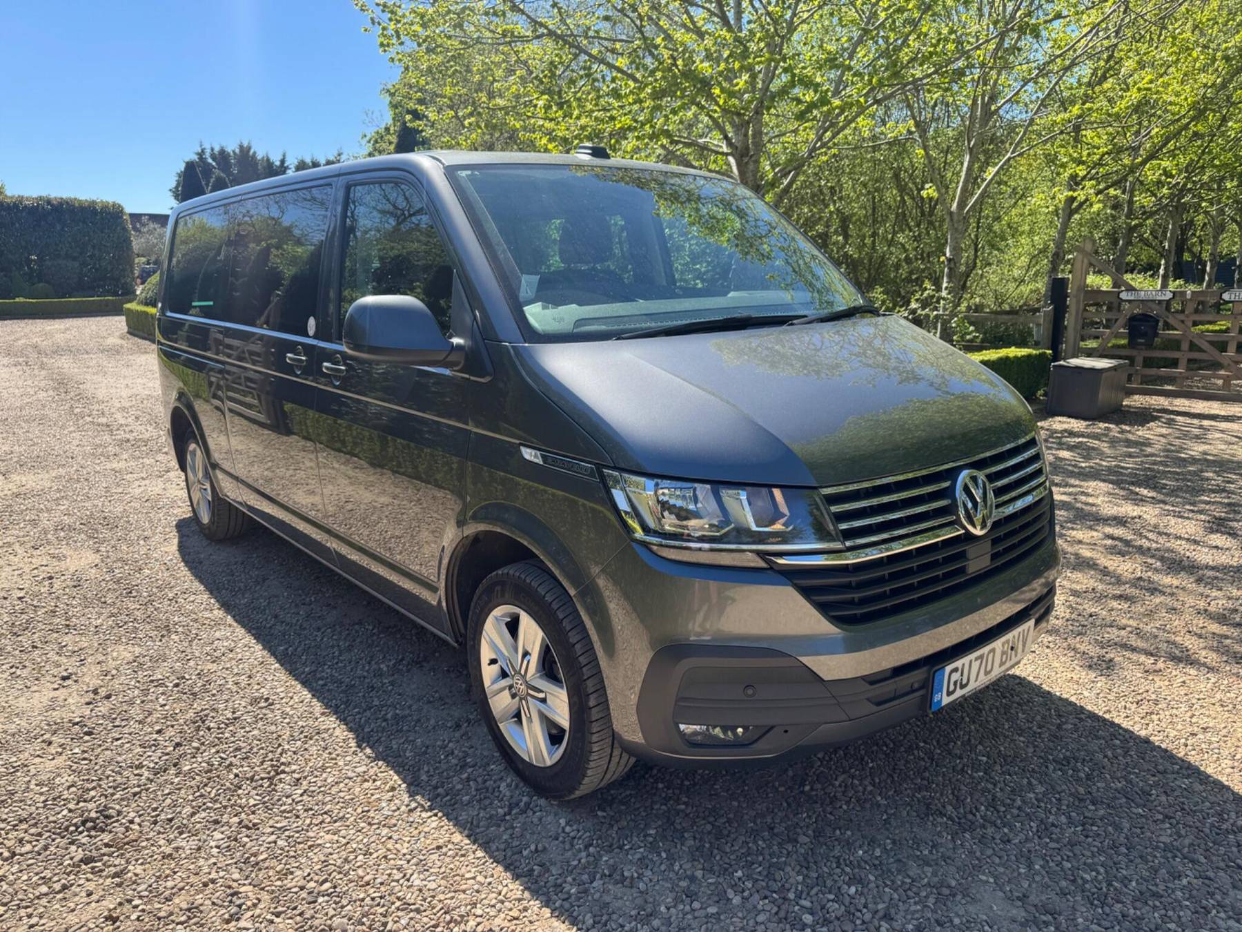 A grey 2020 Volkswagen Caravelle Executive DSG from the front three-quarter, parked on the gravel drive at Rushgreen Farm with trees and a five-bar gate behind