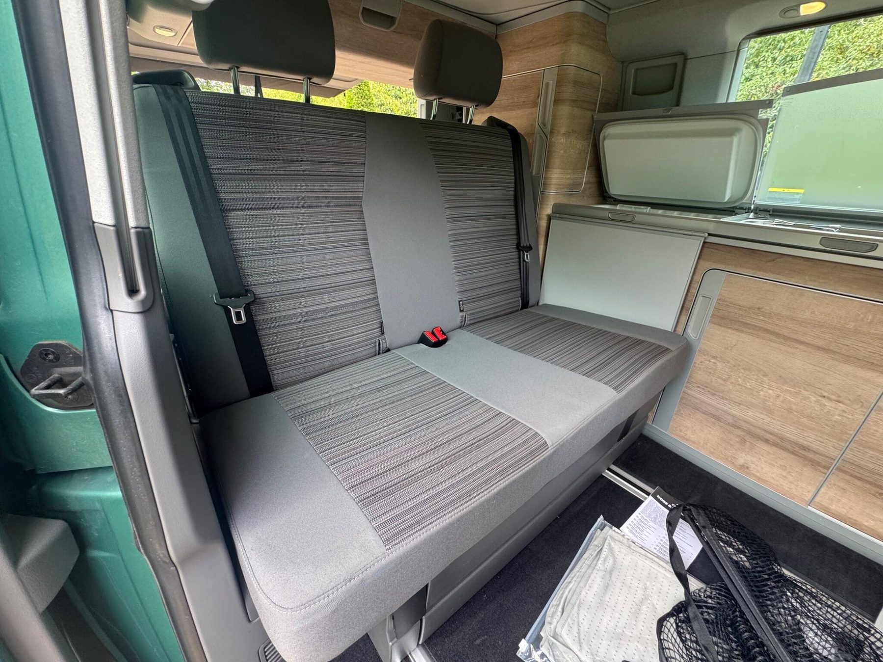 Inside a Volkswagen California Coast: the rear bench seat and kitchen unit in bright oak, with the sliding door open to the trees